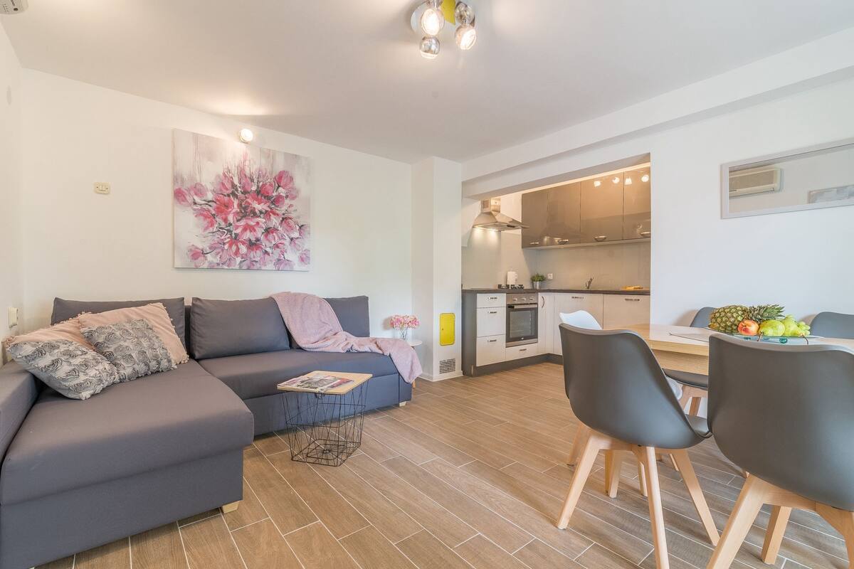 A modern living room with a sofa, a dining table with chairs, a kitchen in the background, and a floral painting on the wall.