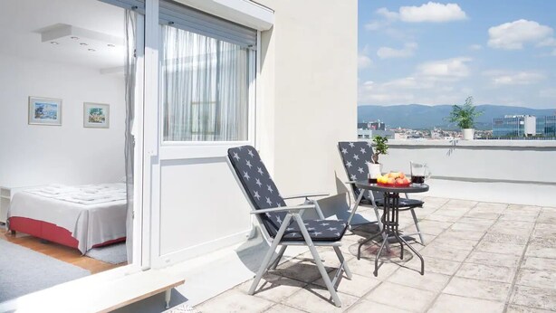 A balcony with a bed, two chairs, a table with fruit, and a view of the city and mountains. - Apartment Asja (Zagreb)
