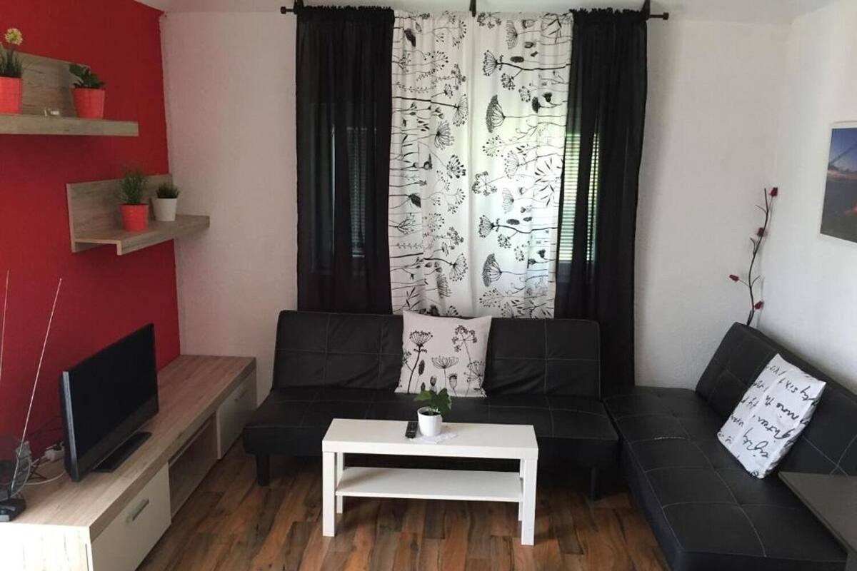 A living room with a black sofa, a white coffee table, and a TV on a wooden stand. There are two red shelves with potted plants.