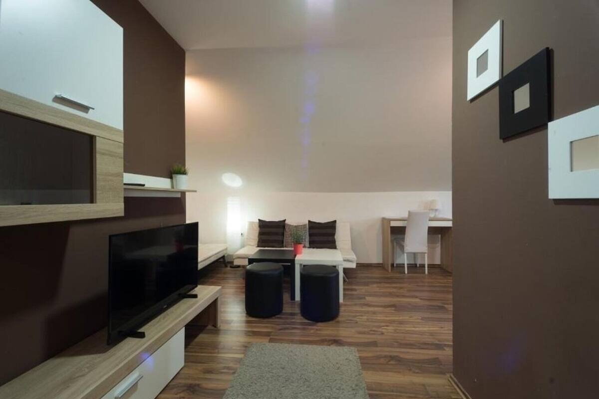 A modern living room with a flat-screen TV, a sofa, a dining table with chairs, and a small kitchen area.