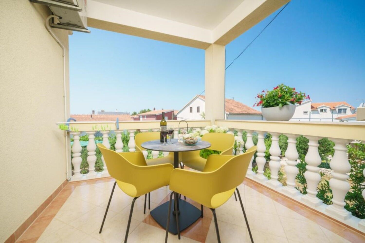 Un balcon avec des chaises jaunes et une table, une plante en pot, et une vue dégagée sur les bâtiments en arrière-plan.
