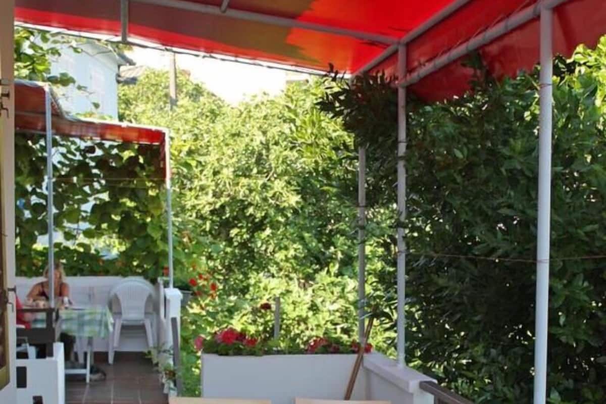 A covered outdoor seating area with a table and chairs, surrounded by greenery.
