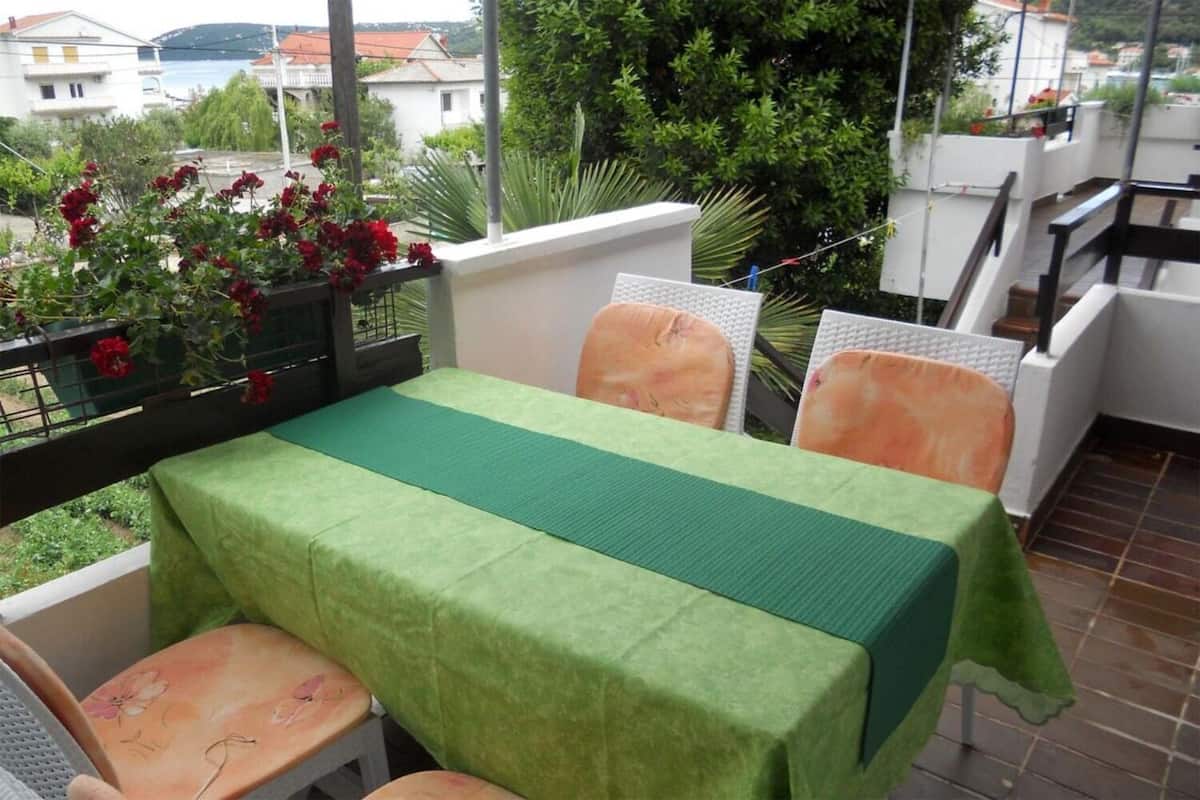 A balcony with a table covered in a green tablecloth, two chairs, and a potted plant with red flowers.