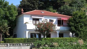 Una casa bianca a due piani con tetto in tegole rosse, un balcone e un giardino con un albero e degli arbusti.