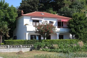 Una casa blanca de dos pisos con techo de tejas rojas, un balcón y un jardín con un árbol y arbustos.