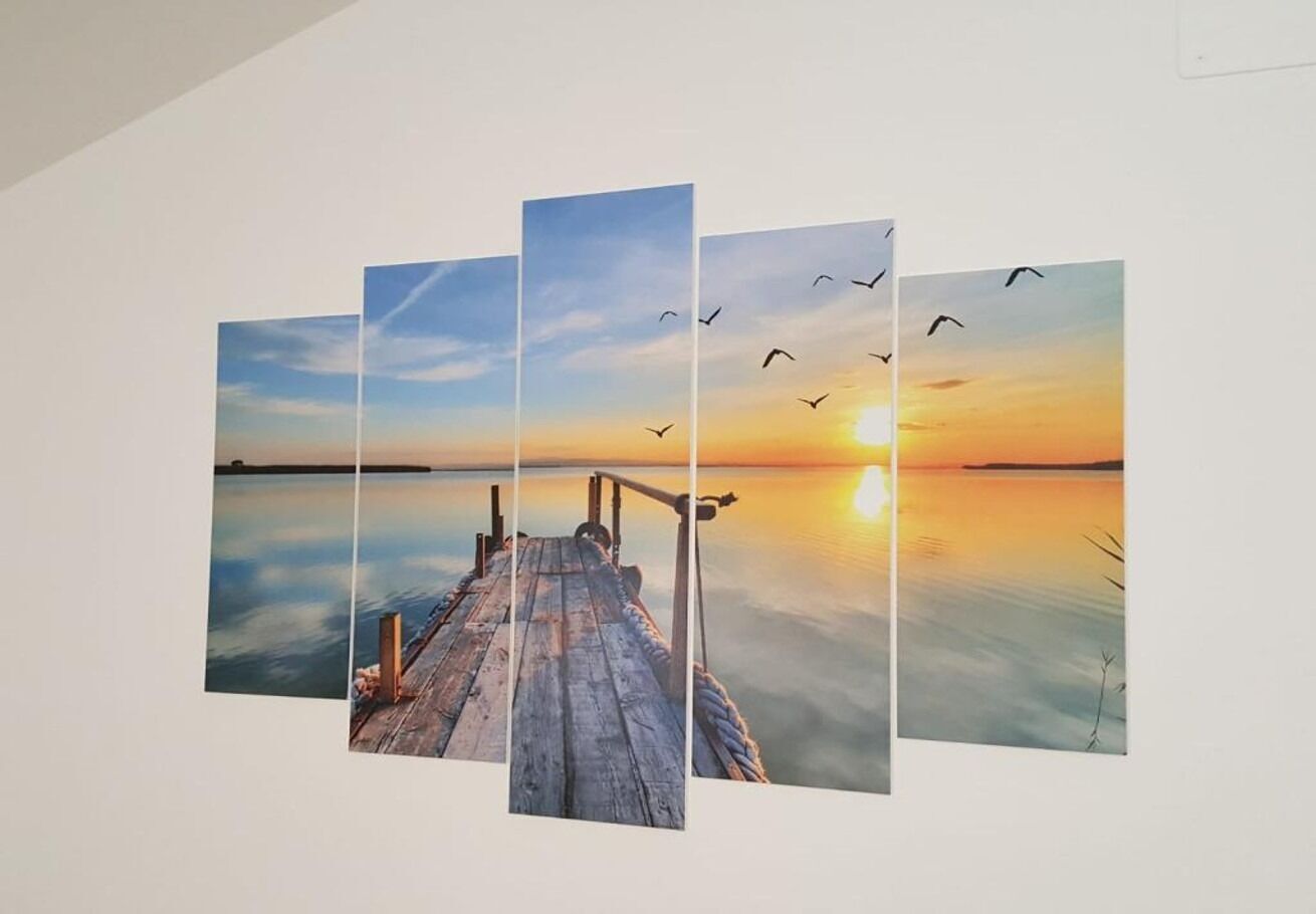 Une œuvre d’art composée de plusieurs panneaux représentant un coucher de soleil sur une étendue d’eau calme, avec une jetée en bois qui s’avance dans le paysage.