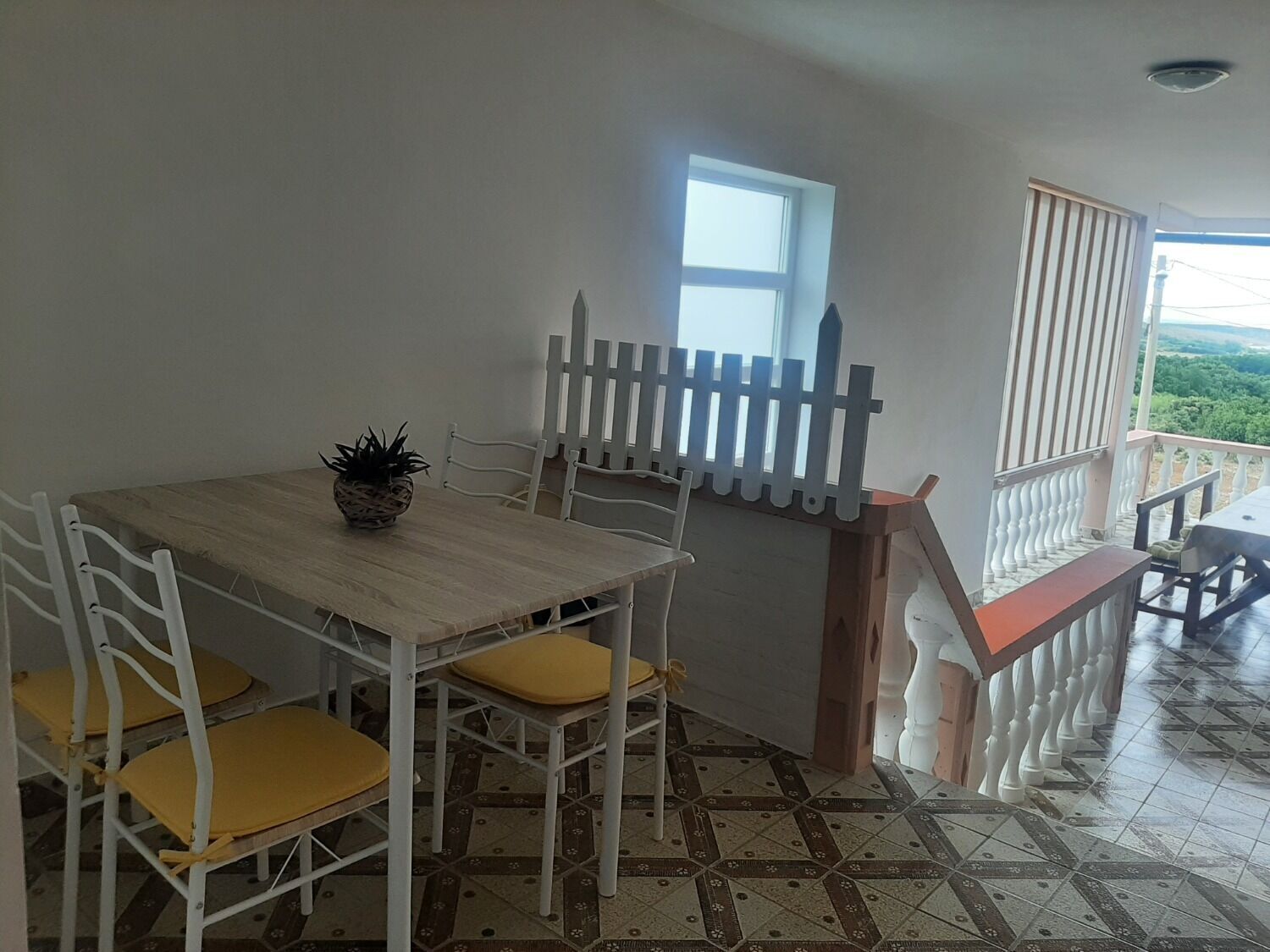 A dining area with a wooden table and white chairs, a potted plant, and a view of greenery through a window.