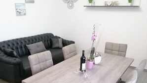 Ein modernes Wohnzimmer mit einem grauen Ecksofa, einem Holzesstisch mit sechs Stühlen und minimalistischer Einrichtung, darunter Wandbilder und ein Regal mit Pflanzen.