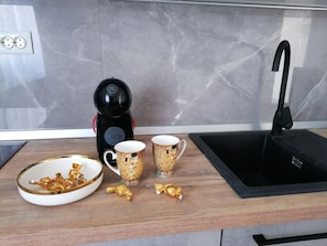 A kitchen counter with a coffee maker, two patterned mugs, a bowl of gold-wrapped chocolates, and a black sink. - Apartment Lilac (Trogir)