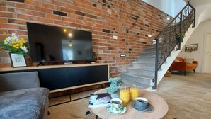 Ein Wohnzimmer mit Backsteinwand, einem Flachbildfernseher, einer Couch, einem Couchtisch mit Getränken und einer Treppe.