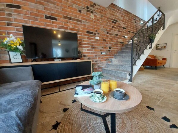 Ein Wohnzimmer mit Backsteinwand, einem Flachbildfernseher, einer Couch, einem Couchtisch mit Getränken und einer Treppe.