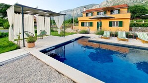 A swimming pool with lounge chairs, a covered seating area, and a yellow building with green shutters. - Villa Vinka (Vrgorac)