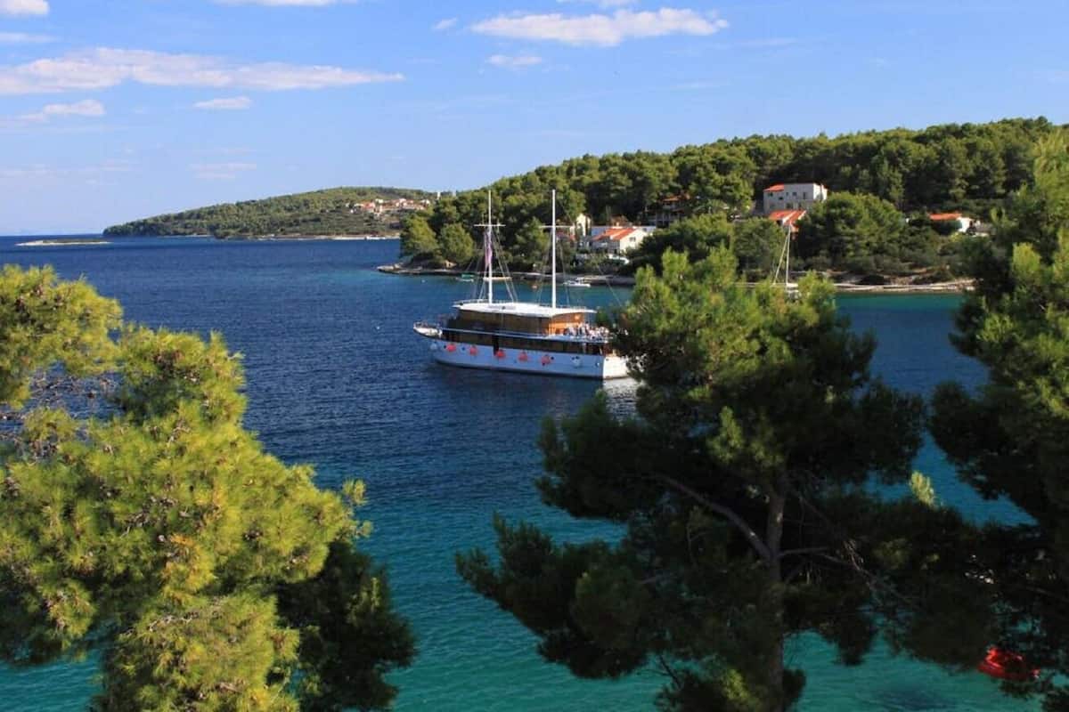 Un paysage côtier avec un bateau sur l’eau, entouré d’arbres et d’une petite colline sur laquelle se trouvent des bâtiments.