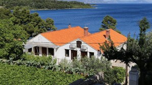 Une maison située sur la côte, avec un toit recouvert de tuiles rouges, entourée d’une végétation luxuriante et offrant une vue sur une mer calme.