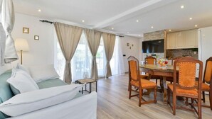 Una camera d'albergo moderna con un letto, un'area pranzo con sedie e un tavolo, una zona cucina e ampie finestre con tende.