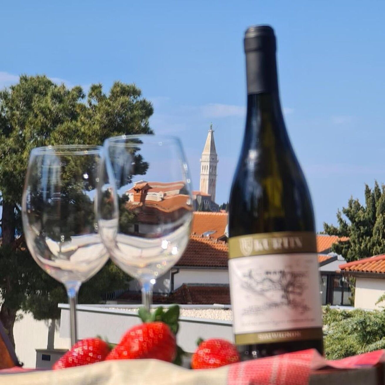 Two wine glasses and a bottle of wine on a table with strawberries, set against a backdrop of a building with a tower.