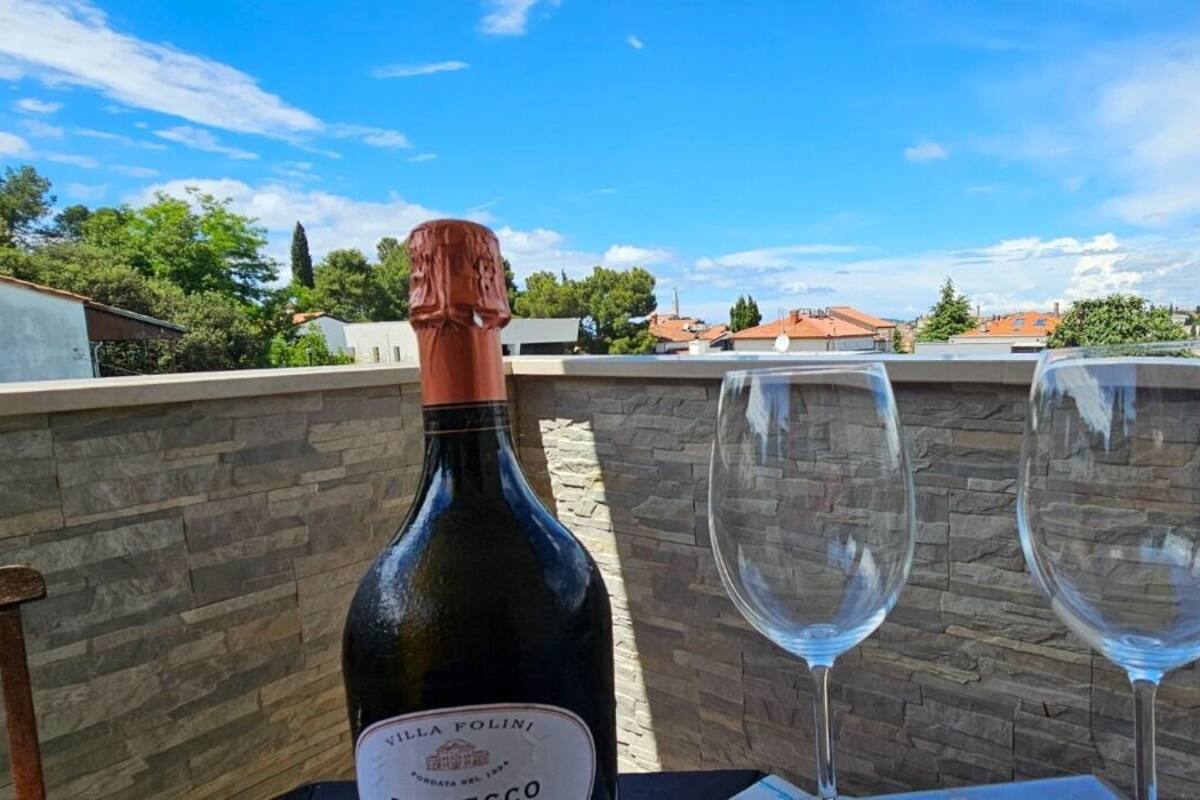 Une bouteille de vin Villa Folini et deux verres à vin vides sont posés sur une table. En arrière-plan, on aperçoit un mur de pierre, ainsi que des maisons et des arbres.