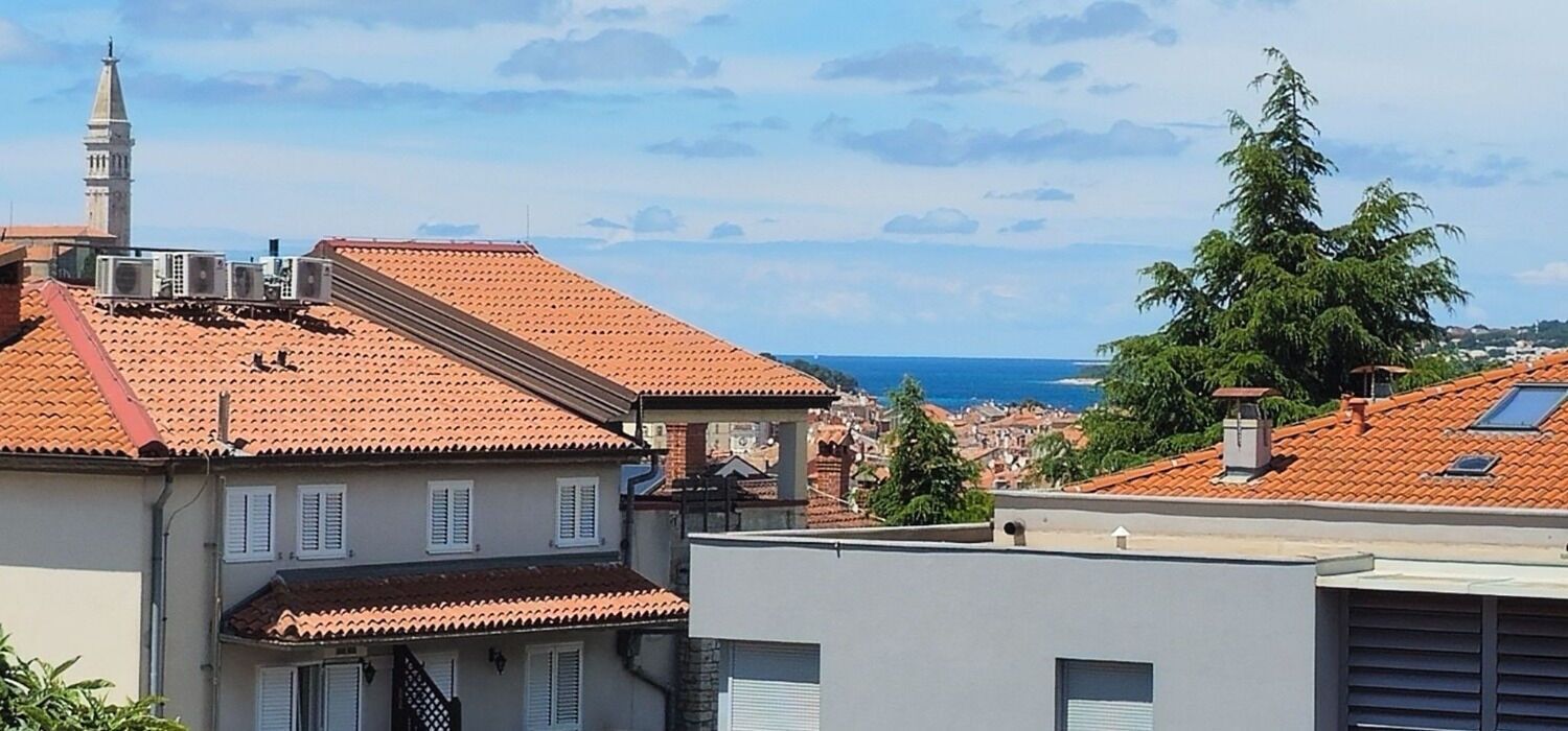 A coastal cityscape with terracotta-roofed buildings, a prominent tower, and a clear view of the sea.