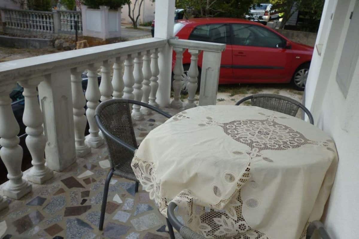 A covered outdoor seating area with a table and chairs, a tiled floor, and a red car in the background.