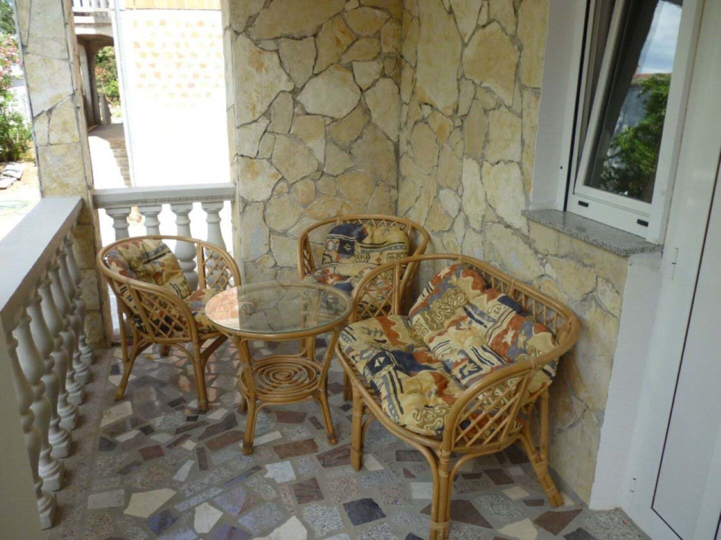 Ein Balkon mit Korbmöbeln, einem Glastisch und einer Steinmauer.