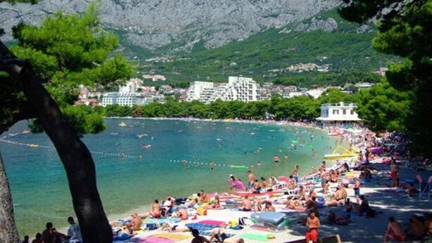 A crowded beach with people sunbathing and swimming, surrounded by mountains and buildings. - Apartments Sunny (Makarska)