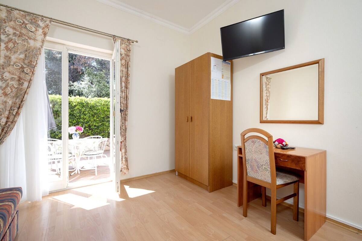 Quarto com guarda-roupa de madeira, escrivaninha com cadeira, televisão de tela plana na parede e porta de vidro deslizante para uma área de estar externa.