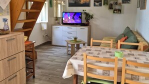 Una sala de estar acogedora con una escalera de madera, un televisor, un comedor con mesa y sillas, y una cocina al fondo.