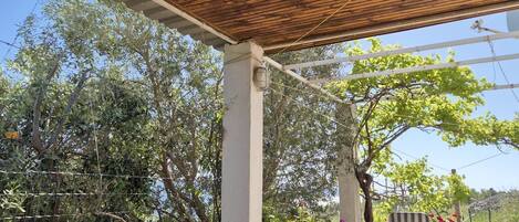 A wooden pergola with a bench underneath, surrounded by greenery and a stone wall.