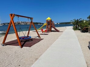 A playground with swings and a slide, a concrete path, and a beach in the background. - Apartments Bibi (Rogoznica)