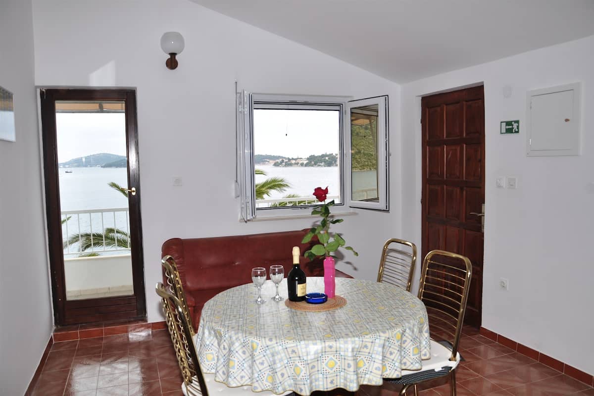 A dining area with a table set for two, a view of the sea through a window, a red rose on the table, and a brown leather couch in the background.