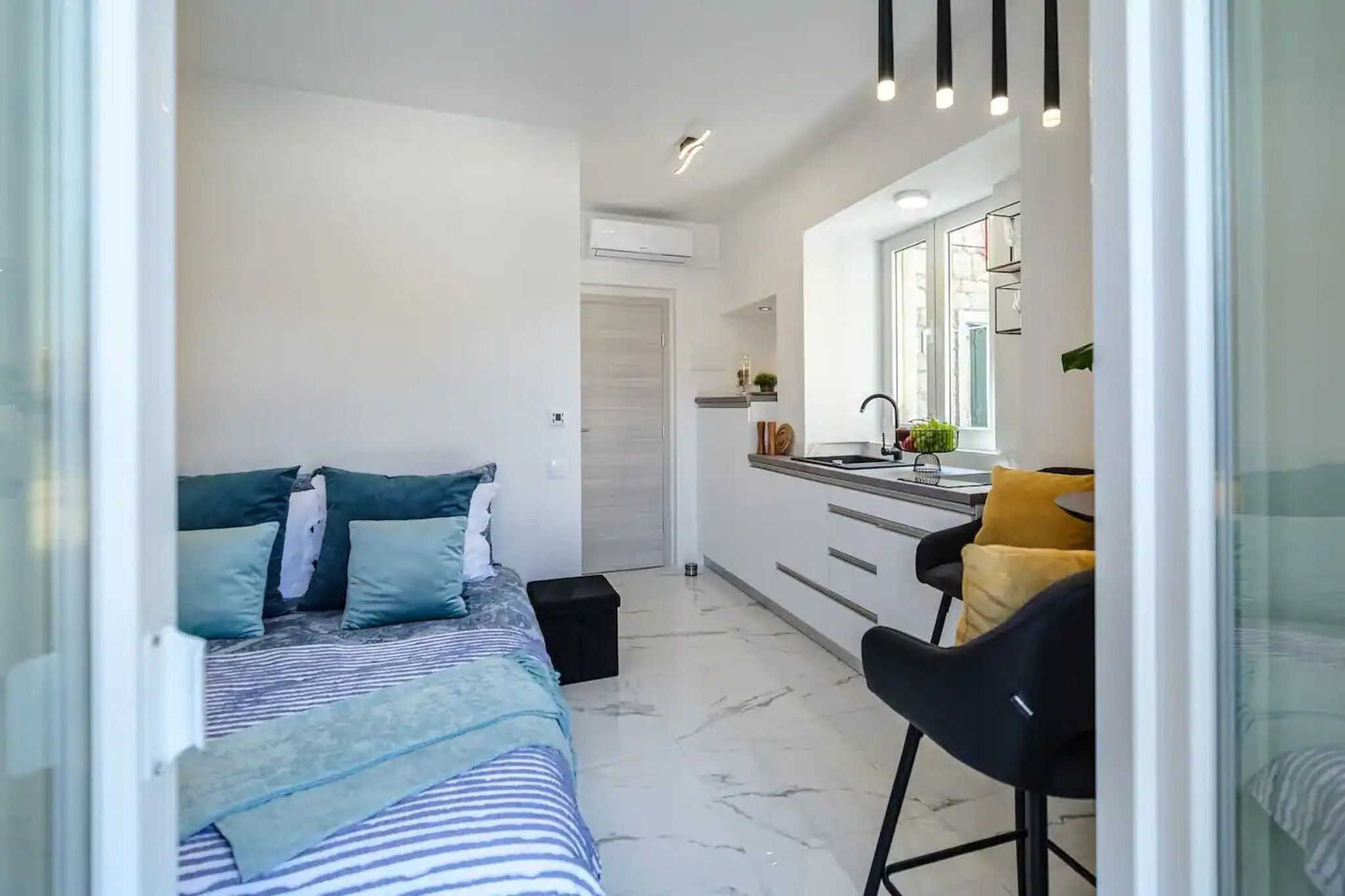 Ein gemütliches Schlafzimmer mit Bett, Nachttisch und einem kleinen Hocker. Eine Küchenzeile ist erkennbar, mit Spüle und Arbeitsfläche.