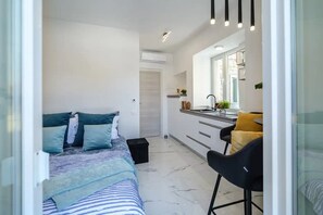 Une chambre à coucher de taille réduite, comprenant un lit, une table de chevet et un petit tabouret. On aperçoit également une petite cuisine avec un évier et un plan de travail.