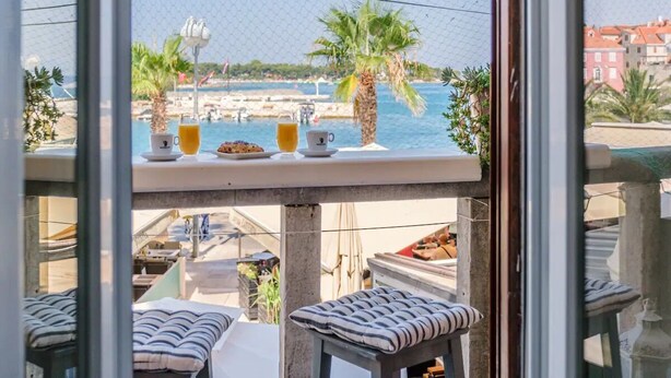 A balcony with a view of the sea and palm trees, a table with breakfast items, and striped cushions. - Apartment Vin (Supetar)