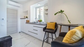 A modern kitchen with white cabinets, a sink, and a window. There is a bar stool with a yellow cushion and a potted plant on the counter. - Apartment Vin (Supetar)