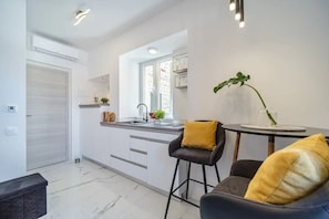 A modern kitchen with white cabinets, a sink, and a window. There is a bar stool with a yellow cushion and a potted plant on the counter. - Apartment Vin (Supetar)