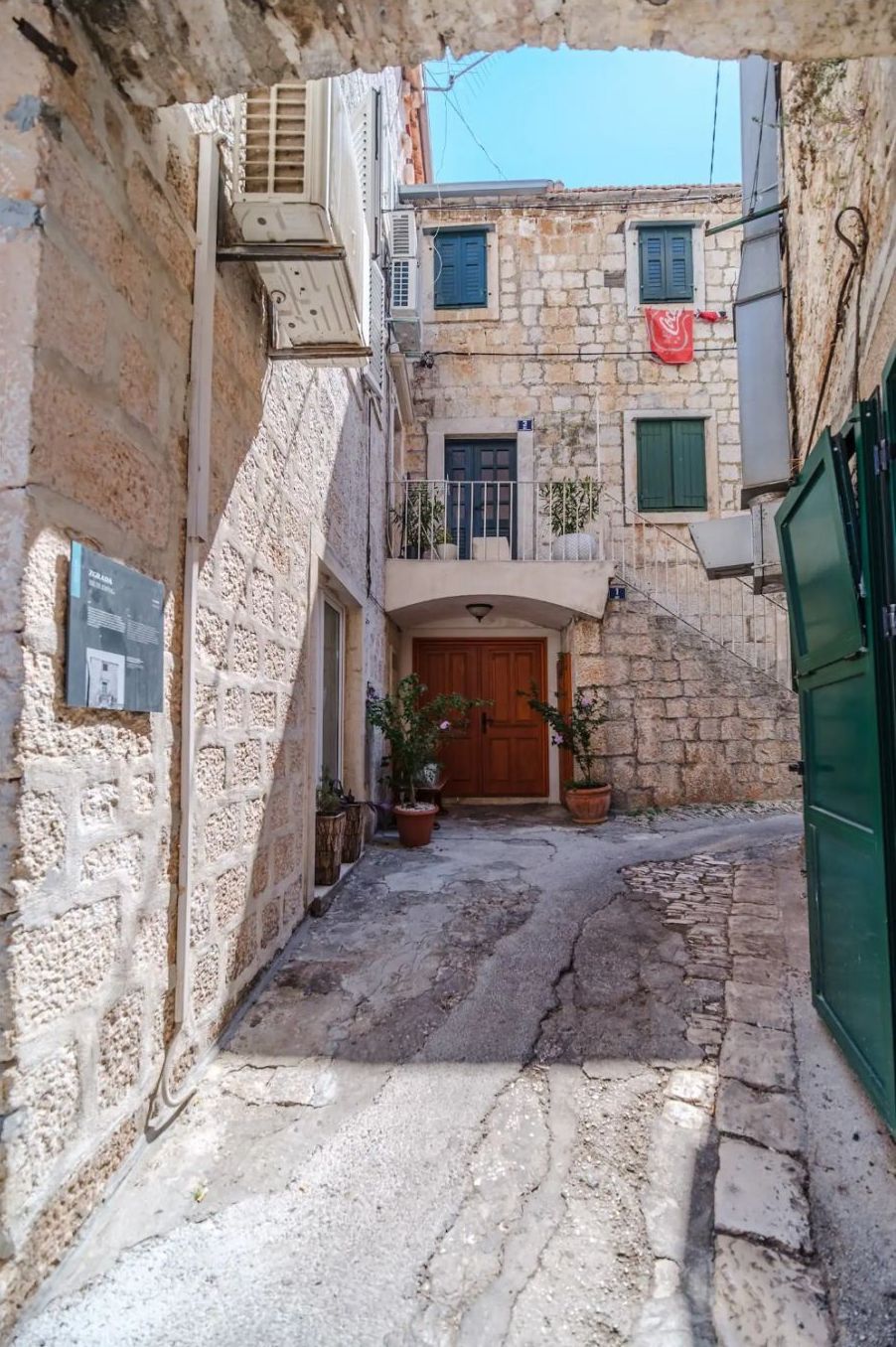 A narrow stone alleyway with a wooden door at the end, flanked by buildings with balconies and potted plants.
