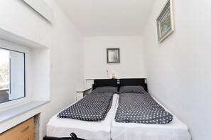 Un petit logement composé d’une seule pièce, comprenant deux lits simples, une table de chevet, une commode, une fenêtre et un tableau encadré au mur.