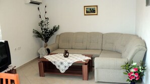 Ein Wohnzimmer mit einem beigefarbenen Sofa, einem Couchtisch aus Holz mit Spitze, einem kleinen orangefarbenen Sessel, einem Fernseher auf einem Schrank, einer Zimmerpflanze und einer Vase mit Blumen.