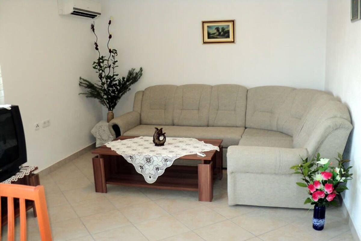 A living room with a beige sofa, a wooden coffee table with a lace tablecloth, a small orange chair, a TV on a stand, a potted plant, and a vase with flowers.