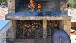 A stone-built outdoor fireplace with a fire burning inside, a chimney, and a stack of firewood beneath. - Apartment Vera (Solta)