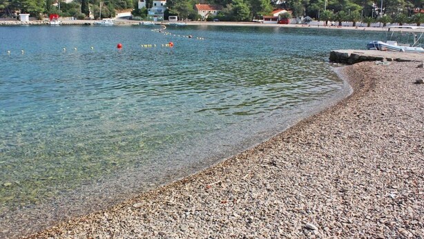 A pebbly beach with clear water, boats, and houses in the background. - Apartment Vera (Solta)