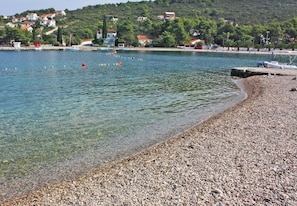 A pebbly beach with clear water, boats, and houses in the background. - Apartment Vera (Solta)