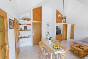Ein Essbereich mit Holztisch, Stühlen und Obstschale. Im Hintergrund befindet sich eine Couch und ein Fernseher.