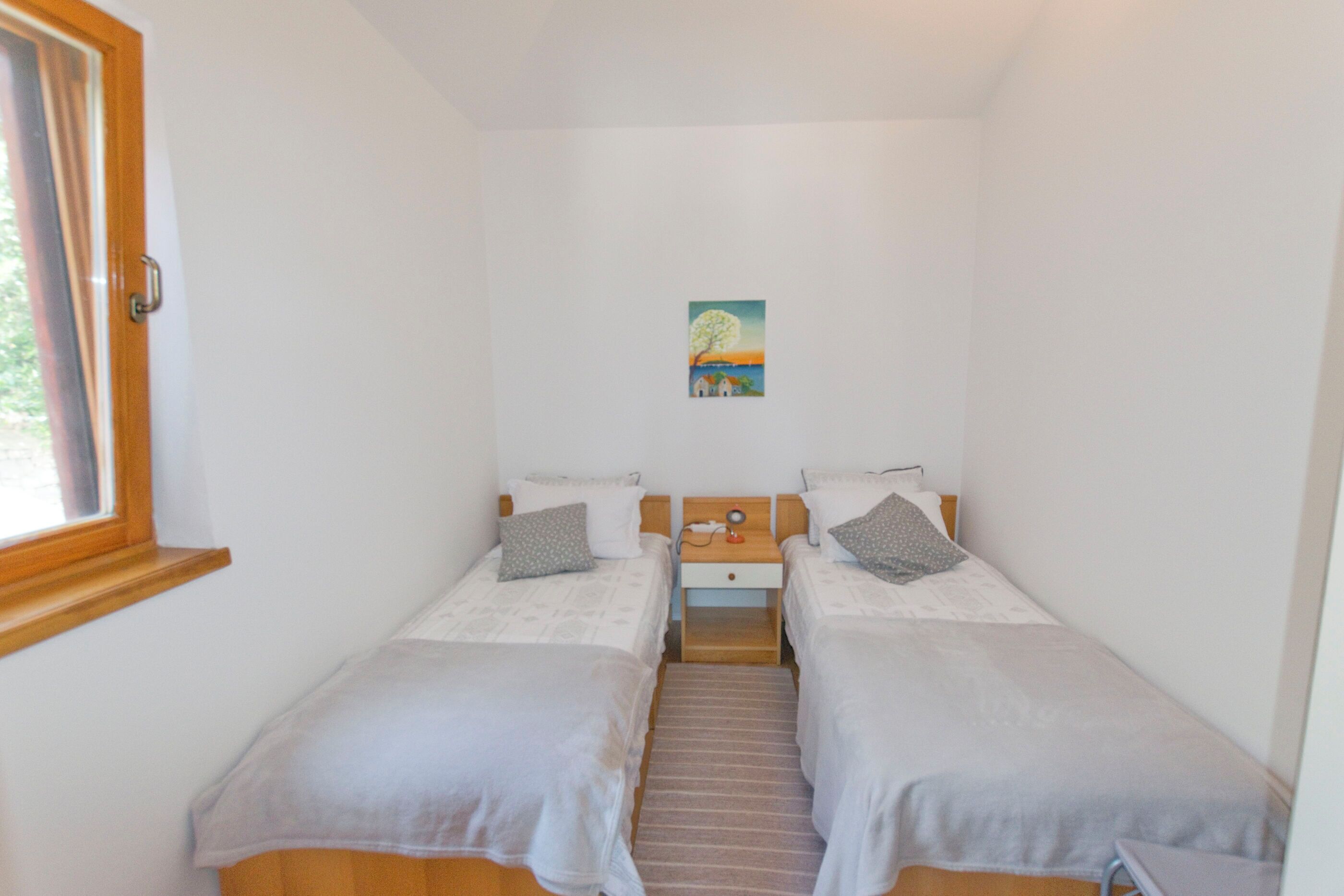 A small, clean room with two single beds, a nightstand, a painting on the wall, and a window with curtains.