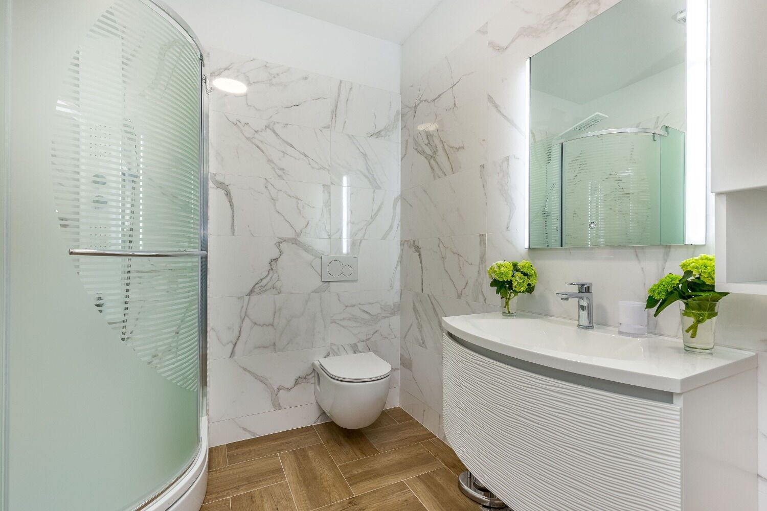 A bathroom with a glass shower enclosure, a toilet, a sink with two flower vases, and a mirror above the sink.