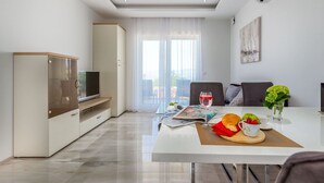 A modern living room with a TV, a dining table with a magazine, a glass of wine, and a fruit basket. - Apartments Igor (Kostrena)