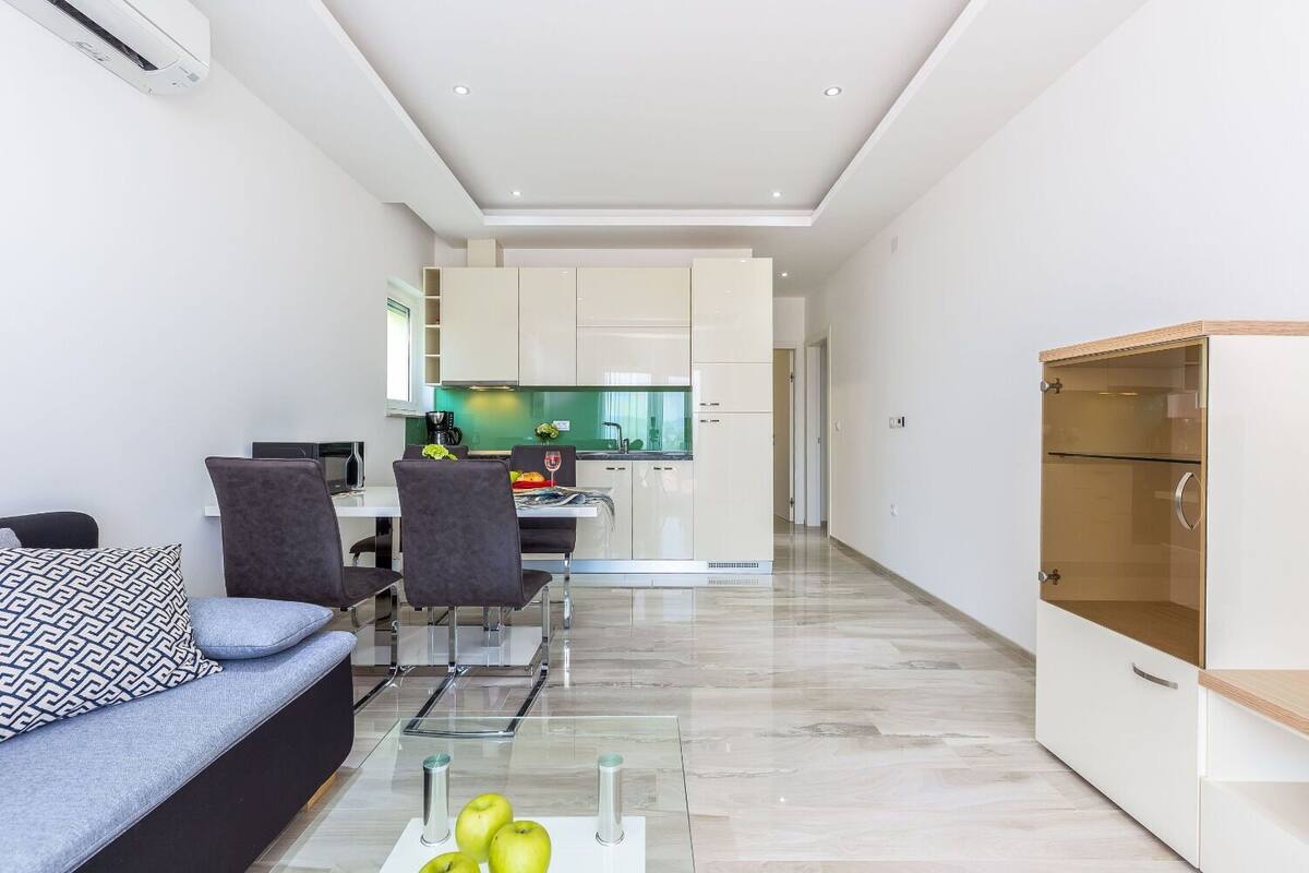 Una sala de estar moderna con un sofá, sillas, una mesa de centro de vidrio y una cocina al fondo.