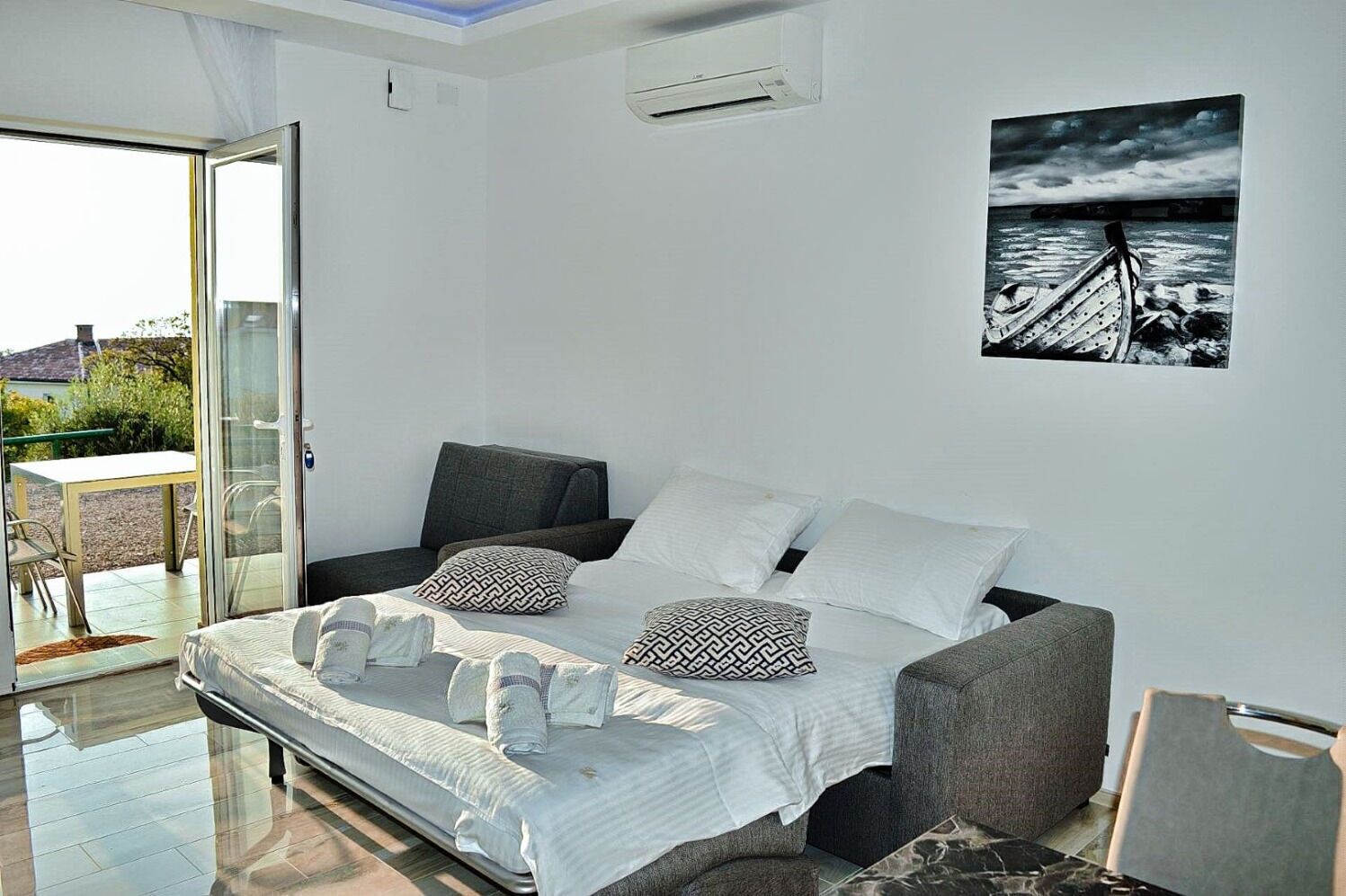 A bedroom with a bed, a grey sofa, a painting of a boat on water, and a sliding glass door leading to an outdoor area.