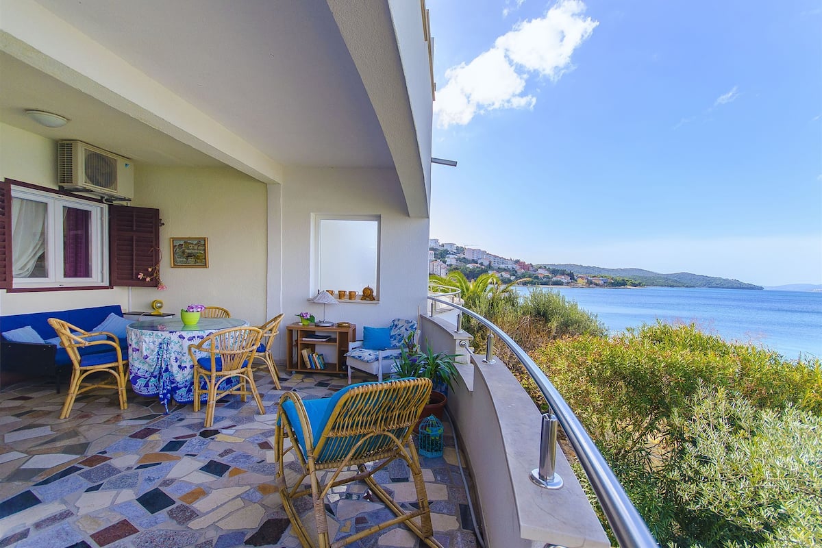 Un balcón con mobiliario de mimbre, una mesa con mantel azul y vista al mar.