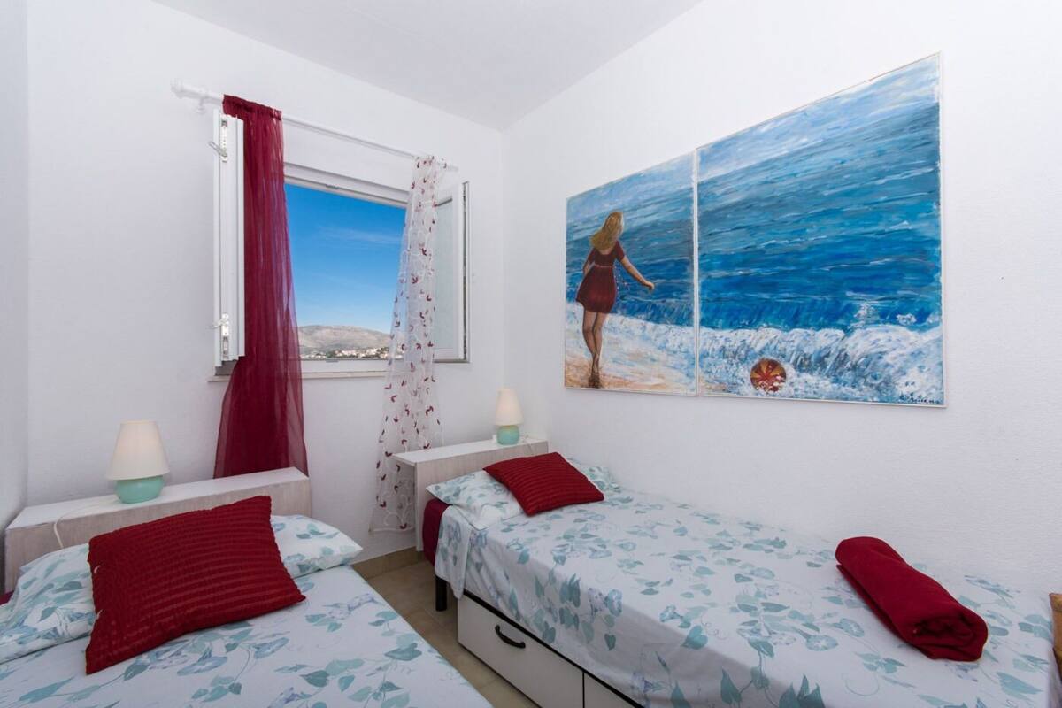 Un dormitorio con dos camas, una ventana con vistas al mar y un cuadro de una mujer junto al mar.